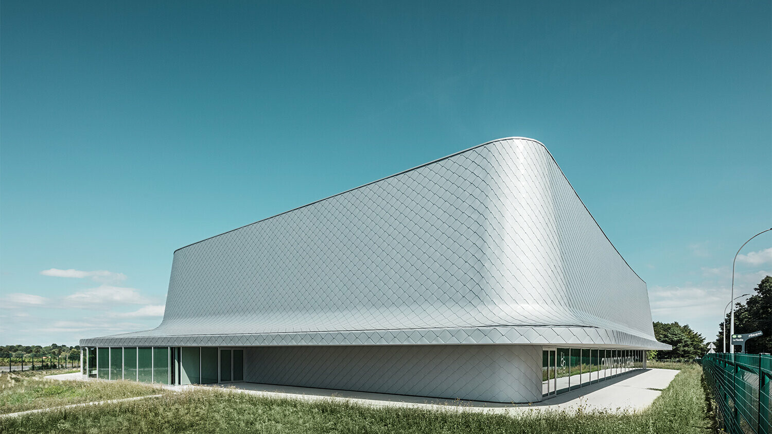 Seitenansicht der Halle, umgeben von der Wiese und einem umliegenden Weg. Die Verglasung ist teilweise sichtbar.