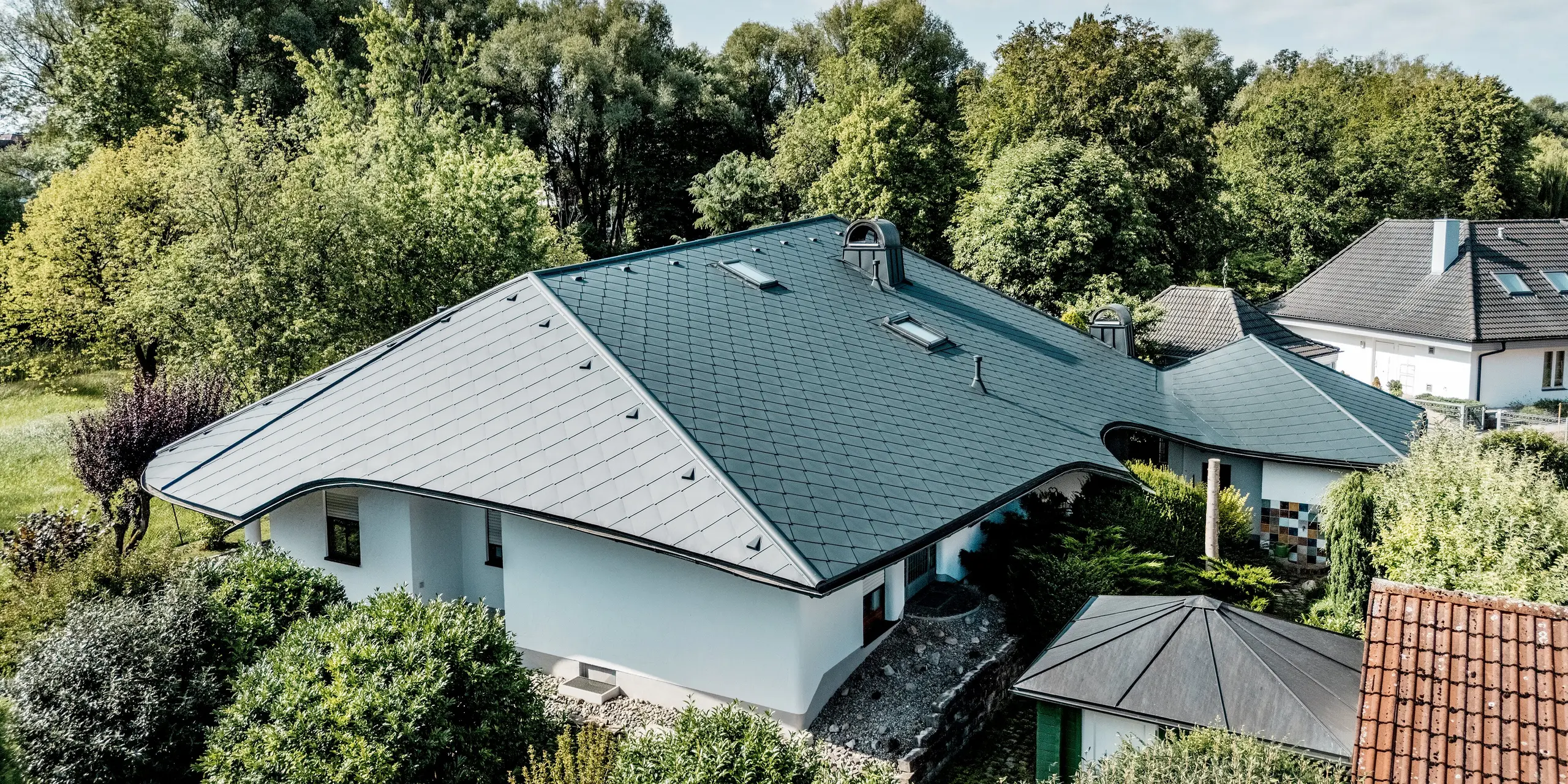 Luftaufnahme des sanierten Einfamilienhauses in Bobingen: Die großzügige Dachlandschaft mit PREFA Dachraute 44 × 44 in P.10 Anthrazit fällt durch geschwungene Trauflinien, markante Dachaufbauten und mehrere Dachfenster ins Auge. Das leichte, robuste Aluminium verbindet hohe Gestaltungsfreiheit mit langlebigem, witterungsbeständigem Schutz.