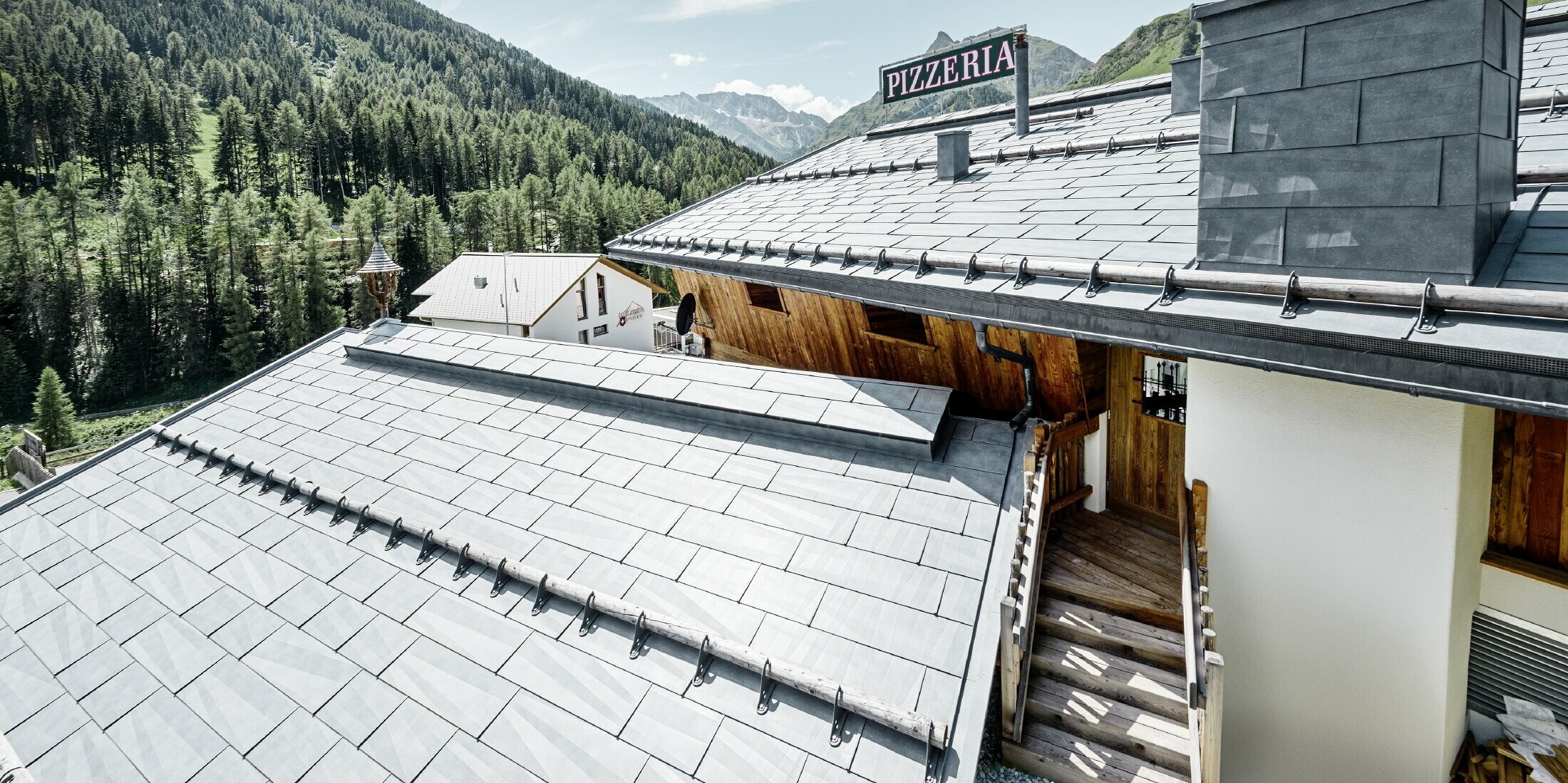 Restaurant Almrausch mit Bergen im Hintergrund und einem PREFA Dach mit dem Dachpaneel FX.12 in steingrau und Gebirgsschneestützen