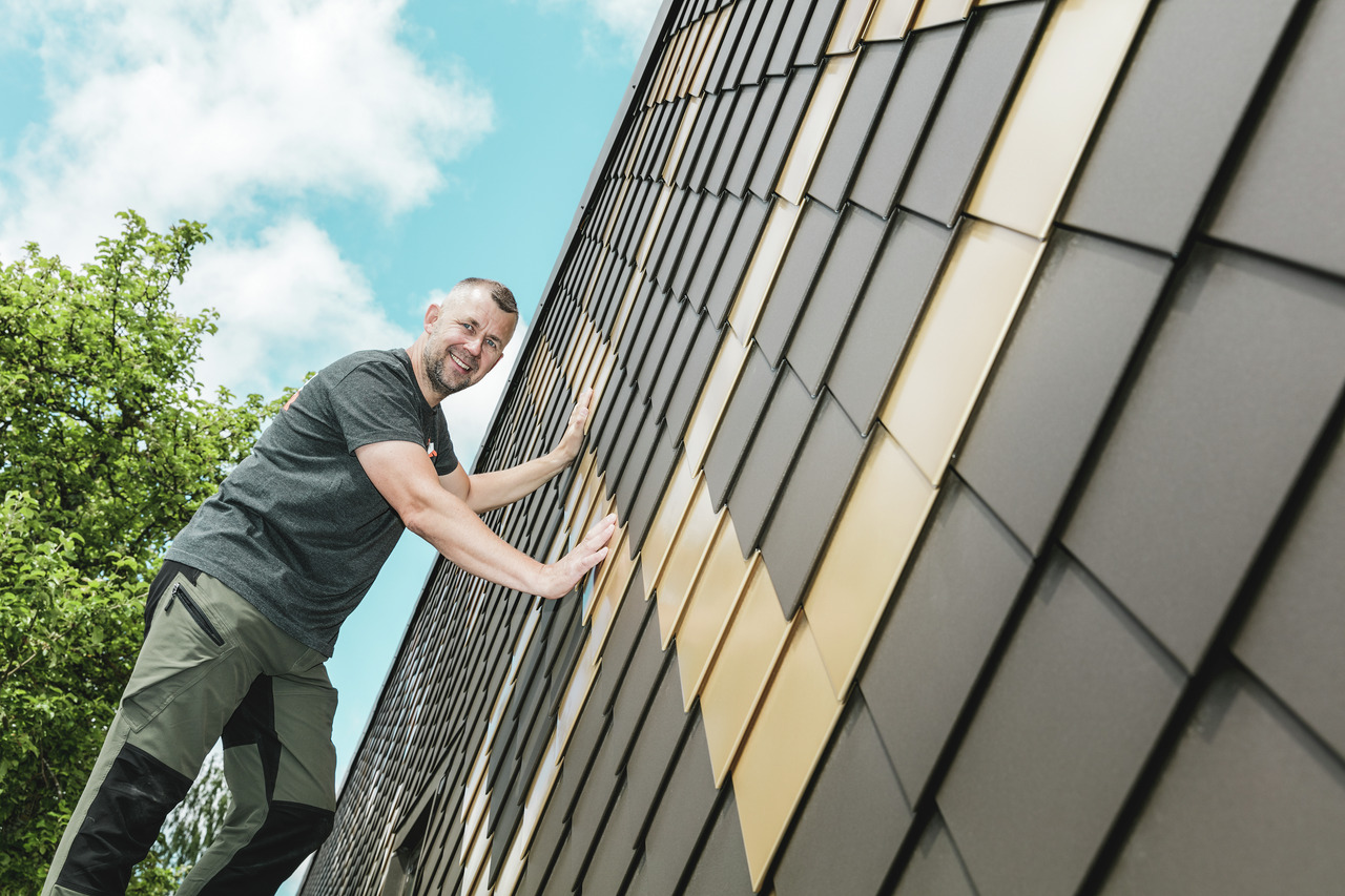 Artisan PREFA portant un t-shirt gris sur une façade en losanges PREFA couleurs brun et or maya.