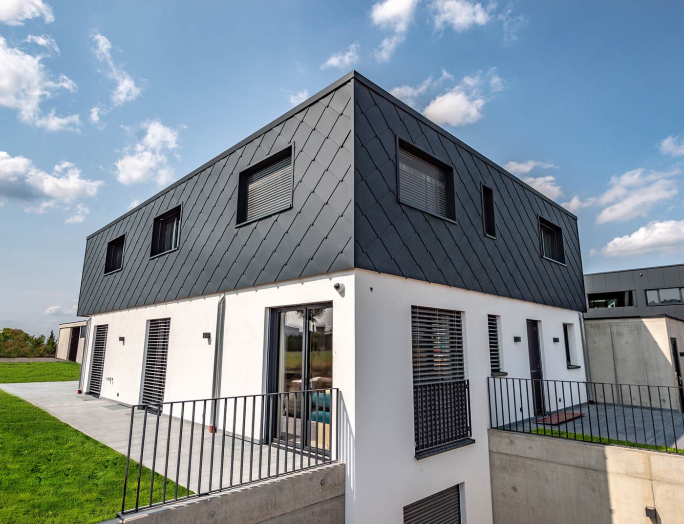 Référence losange de façade PREFA 44 × 44 anthracite Nouvelle construction moderne avec toit plat, façade de l’étage supérieur habillée de losanges de façade en aluminium PREFA 44 × 44 couleur anthracite. La partie inférieure de la façade est revêtue de crépi blanc.