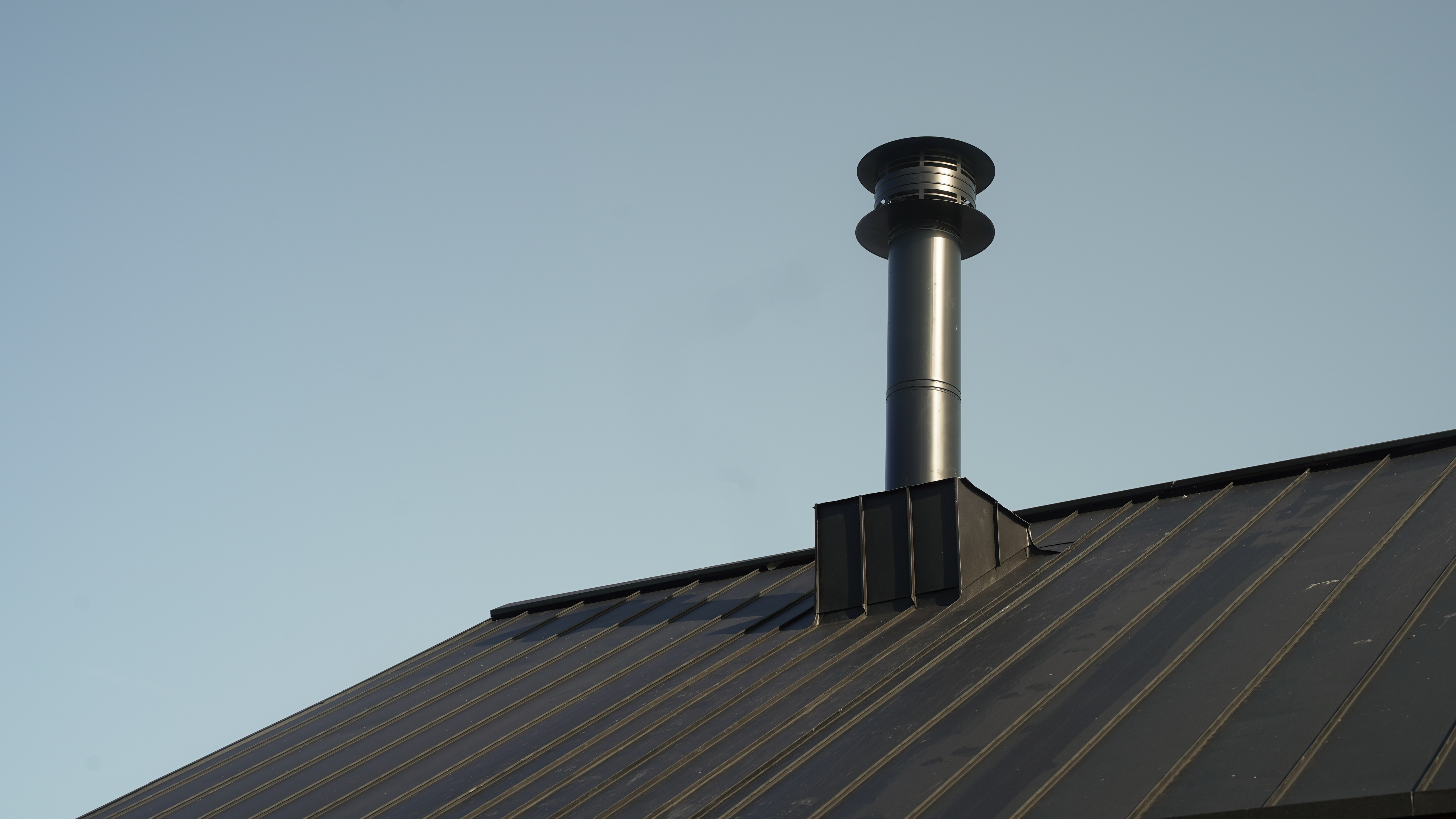 Detailaufnahme eines PREFALZ Dachsystems in P.10 Schwarz am Gebäudekomplex der Golfanlage "Golf it!" in Glasgow, Schottland, mit einem hervorstechenden, modernen Kamin. Der Kamin erhebt sich elegant über die präzise verlegten, dunklen Stehfalzbahnen, die für ihre Langlebigkeit und Robustheit bekannt sind. Dieses Bild unterstreicht die Kombination von Funktionalität und Design, die PREFA Aluminiumprodukte bieten.